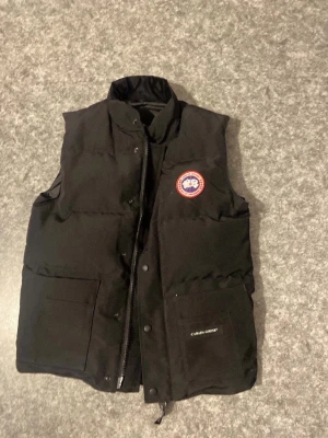 Svart dunväst från Canada Goose - Snygg svart dunväst från Canada Goose med klassisk logotyp på bröstet och två stora fickor framtill. Västen har hög krage, dragkedja och tryckknappar. Perfekt för lager-på-lager och håller dig varm under kyliga dagar.