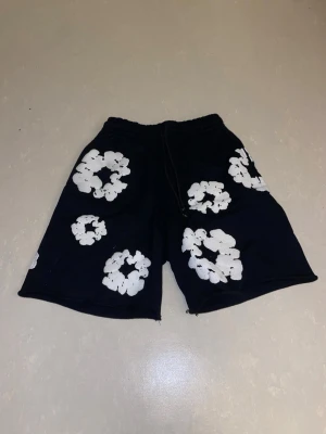 Demin Tears shorts  - Säljer ett par svarta shorts med vita blommönster och snörning i midjan. Shortsen har en avslappnad passform och elastisk midja för extra komfort. Perfekta för sommardagar när du vill ha en cool och unik stil.