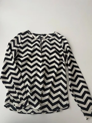 Svartvit zigzag-blus från Lindex - Modellen har rund halsringning med liten knappdetalj framtill och långa ärmar. Materialet är lätt strukturerat och ger en cool look till outfiten.