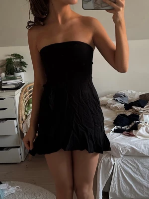 Svart axelbandslös miniklänning - Säljer en svart axelbandslös miniklänning med figurnära passform och lätt utställd kjol. Klänningen har en enkel och stilren design, perfekt för dig som gillar minimalistiskt mode. Materialet är mjukt och följsamt.