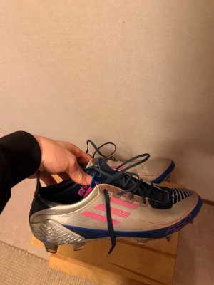 Adidas F50 ”ucl” limited edition  - Säljer ett par Adidas F50 fotbollsskor i silver med blå och rosa detaljer. Skorna har snörning, låg profil och mönstrad ovandel för bättre bollkontroll. Yttersulan är designad för spel på gräsplaner och skon har en sportig, modern look.