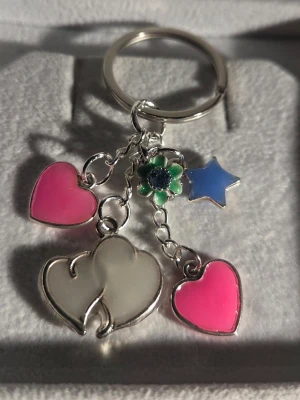 Nyckelring med hjärtan och stjärna - Lyser i mörker - Söt handgjord nyckelring med två rosa hjärtan, en blå stjärna, en vit blomma och två vita hjärtan i metall. Hjärtanen & stjärnan lyser i mörkret och ger en cool effekt. Perfekt att hänga på väskan eller nycklarna för en personlig touch.
