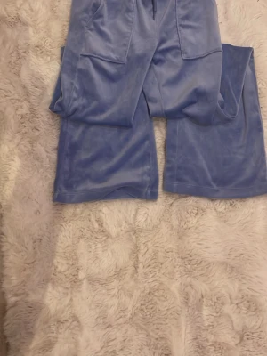 Blå velour mjukisbyxor med utsvängda ben - Säljer ett par ljusblå mjukisbyxor i mjuk velour med utsvängda ben och stora fickor framtill. Byxorna har resår i midjan med dragsko för skön passform. Perfekta för en chill och trendig look.
