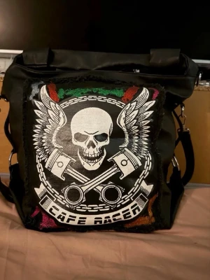 Svart axelväska med Café Racer-tryck - Cool svart axelväska i konstläder med stort patch framtill som har ett Café Racer-motiv med dödskalle, vingar och kolvar. Väskan har justerbar axelrem, silverfärgade metalldetaljer och en fyrkantig form. Perfekt för dig som gillar bikerstil och vill sticka ut.