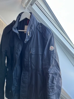 Mörkblå vindjacka från Moncler - Snygg mörkblå vindjacka från Moncler med hög krage, dragkedja och bröstficka med lock. Jackan har Moncler-logga på ärmen och axelklaffar för en sportig look. Tillverkad i lätt, glansigt material som står emot vind och passar perfekt till streetwear. 