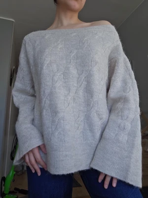 Ljusbeige off shoulder stickad tröja - Mysig ljusbeige stickad tröja från Gina Tricot med bred off shoulder-ringning och kabelstickat mönster. Tröjan har långa, vida ärmar och ribbade muddar. Perfekt för lager på lager och chill stil.