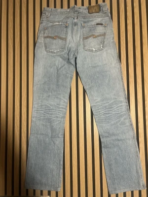 Ljusblå raka jeans från Nudie - Säljer ett par ljusblå jeans från Nudie med raka ben och klassisk femficksmodell. Jeansen har snygga slitningar och kontrastsömmar på bakfickorna. Tillverkade i bomull för en skön och avslappnad känsla. Väldigt populära jeans för ett billigt skick.