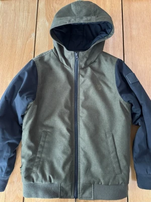 Grön och svart jacka från Element - Stilren jacka från Element i grönt med svarta ärmar och huva. Jackan har dragkedja framtill, två fickor och en patch med Element-logga på ena ärmen. Perfekt för höst och vår, med normal passform och lång ärm. Slitstarkt tyg. 