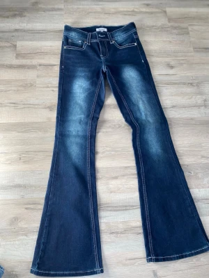 Mörkblå bootcut jeans med kontrastsömmar - Säljer ett par mörkblå bootcut jeans med snygga kontrastsömmar och slitningar framtill. Jeansen har två framfickor och två bakfickor med lock och knappdetaljer. Perfekta för dig som gillar retrovibbar och vill ha ett par jeans med lite vidare ben.