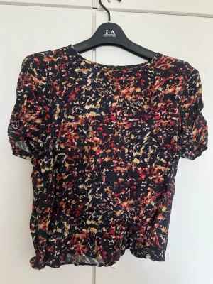 Mönstrad blus med öppning i ryggen från Zara - Säljer en färgglad blus med abstrakt mönster i rött, gult, vitt och svart. Blusen har rund halsringning, korta ärmar och en snygg öppning i ryggen. Materialet känns lätt och luftigt, perfekt för varmare dagar. Passar dig som gillar unika detaljer och färg.