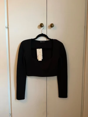 Svart croppad topp  - Trendig svart croppad topp från Zara med lång ärm, axelvaddar och fyrkantig urringning. Toppen är tight i modellen och tillverkad i ett stretchigt syntetmaterial som sitter snyggt på kroppen. Perfekt att styla med höga jeans eller kjol.