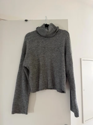 Grå polotröja från H&M - Mysig grå stickad polotröja från H&M i ribbat material. Tröjan har långa ärmar, hög polokrage och en något croppad passform. Perfekt för kalla dagar och enkel att matcha med jeans eller kjol.