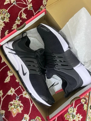 Nike svart mesh sneaker - Snygga svarta Nike sneakers med vit sula och mesh-överdel för bra ventilation. Skon har en låg profil, plastdetaljer vid snörningen och klassisk Nike-logga på sidan. Perfekt för dig som gillar sportig och stilren look.