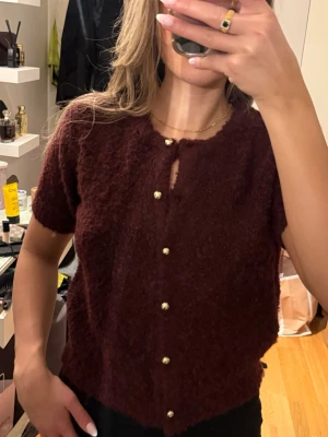 Bordeaux kortärmad kofta med pärlknappar - Mysig bordeauxfärgad kofta med korta ärmar och dekorativa pärlknappar framtill. Koftan har en fluffig, mjuk textur och rund halsringning. Perfekt för att addera lite lyxig känsla till din outfit.