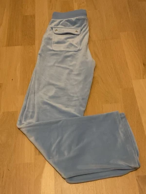 Äkta ljusblå velourbyxor Juicy Couture - Säljer ett par ljusblå mjukisbyxor från Juicy Couture i mjuk velour. Byxorna har en bakficka med knappdetalj och klassisk relaxed fit. Perfekta för en chill och trendig look. Nytt, ej använt.