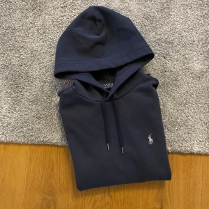 Mörkblå hoodie från Polo Ralph Lauren - Snygg mörkblå hoodie från Polo Ralph Lauren med klassisk huva och dragsko. Har en broderad vit logga på bröstet och en stor magficka framtill. Tillverkad i mjukt bomullsmaterial och har ribbade muddar vid ärmslut och nederkant.