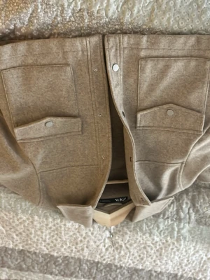 Beige overshirt/kappa  från Zara - Stilren beige overshirt/kappan från Zara med två stora bröstfickor och tryckknappar framtill. Overshirt/kappan har klassisk krage och är gjord i ett mjukt, filtliknande material som ger en avslappnad men trendig look. Perfekt att slänga över en hoodie eller t-shirt. Den är ifrån Zara i storlek S köpt för nått år sen och är använd en gång 💞