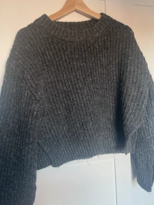 Mörkgrå ribbstickad tröja - Mysig mörkgrå stickad tröja med bred ribbstickning och avslappnad passform. Tröjan har rund halsringning, långa ärmar och ribbade slits vid ärmslut och nederkant. Perfekt för kyliga dagar och enkel att matcha till olika outfits.