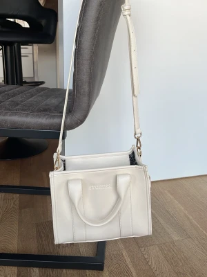 Vit axelväska från & Other Stories - Snygg vit/beige axelväska som är köpt på Ullared i boxig modell. Väskan har justerbar axelrem, två handtag och guldfärgade detaljer. Tillverkad i slätt konstläder med diskret logga framtill. Perfekt för dig som gillar stilrena accessoarer.