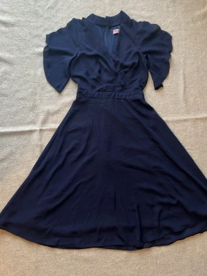 Marinblå omlottklänning med v-ringning - Snygg marinblå klänning från Collectif med omlottliv och v-ringning. Klänningen har korta, lösa ärmar och markerad midja. Kjolen är lång och svepande, vilket ger en elegant siluett. Perfekt för dig som gillar klassisk och stilren design.