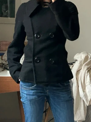 Svart kappa med dubbelknäppning - Stilren svart kappa med bred krage och dubbelknäppning framtill. Figurnära passform som markerar midjan och ger en snygg siluett. Perfekt som vårjacka. Har hål i fickorna men inget som är synligt