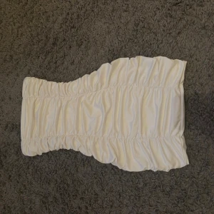 Vit axelbandslös kortklänning - Trendig vit axelbandslös kortklänning med snygga rynkade detaljer. Klänningen har en figurnära passform och är perfekt för dig som vill ha en enkel men stilren look. Tillverkad i ett mjukt material som känns bekvämt mot huden.