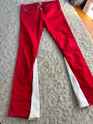 Röda bootcut jeans med vita detaljer - Säljer ett par röda bootcut jeans med vita paneler längs insidan av benen som jag sytt själv. Jeansen har dragkedjor på fickorna både fram och bak samt en klassisk femficksdesign. Materialet är troligtvis bomull med lite stretch för skön passform.