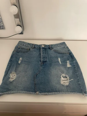 Blå jeanskjol med slitningar - Säljer en blå jeanskjol i kort modell med slitna detaljer och rå kant nertill. Kjolen har klassisk femficksdesign och stängs med knapp och dragkedja framtill. Perfekt för dig som gillar en avslappnad och trendig stil.