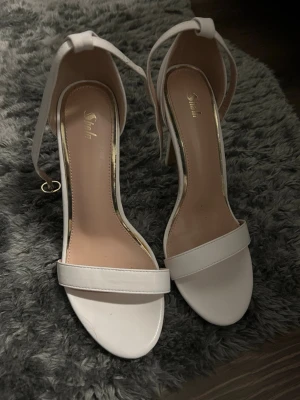 Vita stilettklackar från Shein - Säljer ett par vita pumps från Shein med öppen tå, smal rem över tårna och justerbar ankelrem. Skorna har höga stilettklackar och en stilren, minimalistisk design. Materialet är konstläder med en mjuk innersula för extra komfort. Finns lite märken på skorna men annars fint skick.