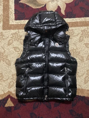Moncler vest NFC - Säljer en svart glansig dunjacka från Moncler med klassisk logotyp på bröstet. Jackan har dragkedja framtill och är fylld med dun för extra värme. Perfekt för kalla vinterdagar och har en modern, puffig look.