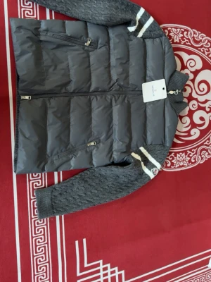 Grå Moncler dunväst med stickade ärmar - Snygg grå dunväst från Moncler med stickade ärmar i kabelstickat mönster och vita ränder. Västen har två dragkedjeförsedda fickor och ribbade muddar. Klassisk Moncler-logga på ärmen och hög krage för extra stilpoäng.