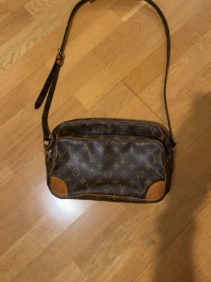 Äkta Louis Vuitton Monogram axelväska - Säljer en klassisk Louis Vuitton axelväska i brunt monogrammönster med detaljer i ljusbrunt läder. Väskan har justerbar axelrem, en framficka med dragkedja och rektangulär form. Perfekt för dig som gillar lyxiga accessoarer och tidlös design.