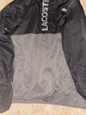 Svart och grå vindjacka från Lacoste - Snygg vindjacka från Lacoste i svart och grått med stor logga på bröstet och den klassiska krokodilen på bröstet. Jackan har dragkedja, två sidofickor och är tillverkad i ett lätt, vindtätt material. Perfekt för en sportig och avslappnad stil.Har byxor med men säljs separat/ Storlek S/M