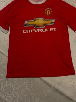 Manchester United Di Maria tröja - Röd Manchester United matchtröja med vitt tryck, Chevrolet-logga på bröstet och Di Maria samt nummer 7 på ryggen. Klassisk rund hals med vit kant och klubbmärke på vänster bröst. Perfekt för fotbollsfans!