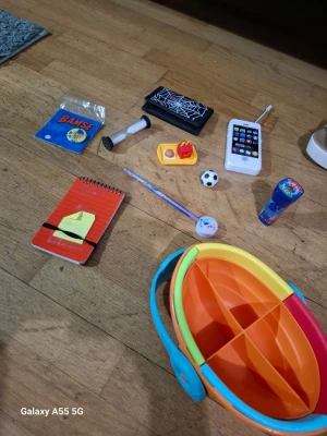 Blandade småprylar och leksaker - Säljer ett paket med blandade småprylar och leksaker, inklusive en orange anteckningsbok, plastbåt i orange och gult, leksaksmobil, timglas, penna, liten fotboll, blå såpbubbelburk och några små tillbehör. Perfekt för lekar, pyssel eller som överraskning.