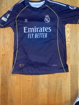Real Madrid Mbappé matchtröja blå - Snygg blå Real Madrid matchtröja med Mbappé och nummer 9 på ryggen. Tröjan har vita och gula detaljer, klubbmärke på bröstet och Emirates Fly Better-tryck. Tillverkad i lätt och ventilerande material, perfekt för fotbollsträning eller match.