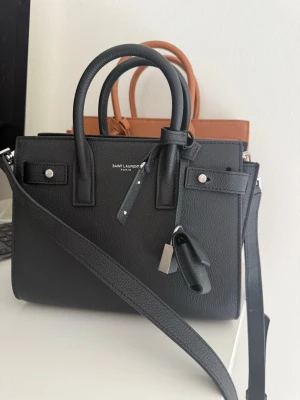 Svart  Sac de Jour väska - Säljer en svart Sac de Jour handväska  i grained läder. Väskan har både handtag och axelrem, silverfärgade metalldetaljer och ett hänglås med logga. Klassisk boxig form och stilren design med diskret logotyp framtill.