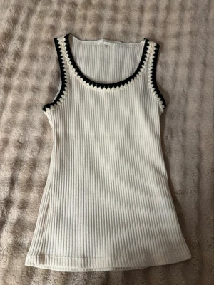 Vit ribbad linnetopp från H&M - Säljer en vit ribbad linnetopp från H&M i storlek XS. Toppen har svarta virkade detaljer runt halsringning och ärmhål, vilket ger en snygg kontrast. Perfekt till jeans eller kjol för en fräsch look.