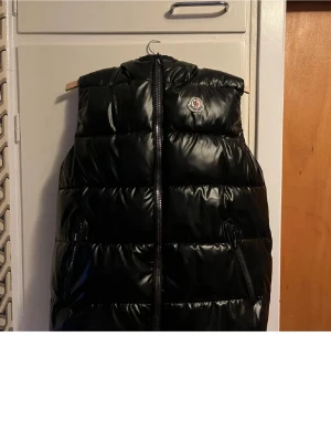 Svart dunväst från Moncler - Snygg svart dunväst från Moncler med glansig finish och hög krage. Västen har två dragkedjefickor framtill och Moncler-logga på bröstet. Perfekt för lager på lager och ger en cool streetwear-look.