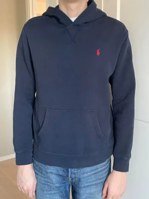 Mörkblå hoodie från Polo Ralph Lauren - Mörkblå hoodie från Polo Ralph Lauren med klassisk röd logga broderad på bröstet. Hoodien har huva, känguruficka och ribbade muddar vid ärmslut och nederkant. Tillverkad i mjukt bomullsmaterial som är skönt att bära. Nypris 1295 kr.