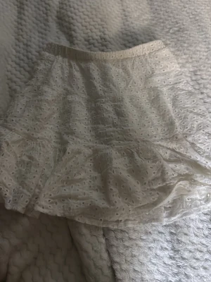 Vit kort kjol med volang  - Supersöt vit kort kjol med volangkant och broderade hål. Kjolen har resår i midjan och är gjord i ett lätt bomullstyg som känns luftigt och somrigt. Perfekt för dig som gillar romantiska detaljer och vill ha en feminin look.