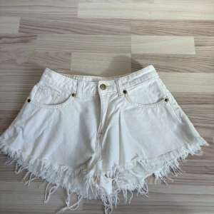 Vita fransiga shorts i bomull - Säljer ett par vita shorts med fransig kant och klassisk femficksdesign. De har hög midja och stängs med knapp och dragkedja. Perfekta för varma dagar och ger en avslappnad look.