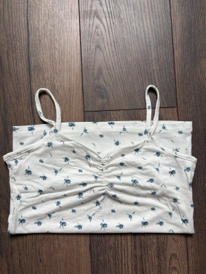 Vit linne med blåbär på - Söt vit linnetopp med tunna axelband och små blåbär över hela tyget. Toppen har rynkdetalj framtill vid bysten och är gjord i mjuk bomull, perfekt för varma dagar. Enkel och stilren design som passar till mycket.