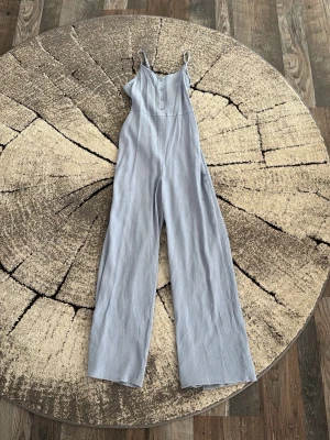 Ljusblå jumpsuit med knappar - Säljer en ljusblå jumpsuit med smala axelband och raka, vida ben. Framtill finns fyra dekorativa knappar och tyget är mjukt och tunt, troligen i viskos eller bomullsblandning. Perfekt för en avslappnad och trendig look.