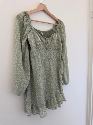 Grön blommig kortklänning med volang - Söt ljusgrön kortklänning med små vita blommor, långa ärmar och volangkant nertill. Klänningen har en omlottliknande detalj på sidan, knyt framtill vid bysten och är tillverkad i ett lätt, luftigt material. Perfekt för sommarens alla tillfällen.