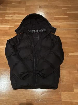 Svart pufferjacka från Tommy Hilfiger - Snygg svart pufferjacka från Tommy Hilfiger med huva och diskret logga på bröstet samt på axeln. Jackan har dragkedja framtill och quiltad design samt två fickor där framme. Perfekt för kalla vinterdagar och riktigt skön att bära. Inga defekter eller liknande. (Priset är inte hugget i sten) Nypris cirka 2300-2500kr. 
