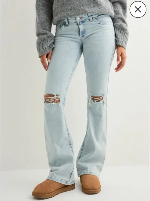 Ljusblå bootcut jeans med slitningar - Helt slutsålda nelly jeans! Säljer ett par ljusblå bootcut jeans med slitningar över knäna och broderade detaljer i rosa på bakfickorna. Jeansen har hög midja och klassisk femficksdesign. Tillverkade i denim med en avslappnad passform och trendig look.