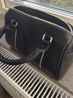 Svart handväska med silverdetaljer - Snygg svart handväska i skinnimitation med två rejäla handtag och silverfärgade dragkedjor. Väskan har en boxig form och flera fack med dragkedja framtill. Perfekt för dig som gillar stilrena accessoarer med lite edge.