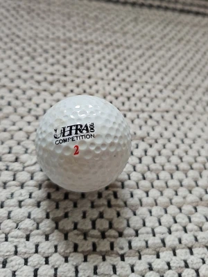 Ultra Competition golfboll - Säljer en vit golfboll från Ultra Competition med text och siffran 2 i rött. Klassisk rund form med dimple-mönster för bättre flyg och kontroll. Perfekt för dig som gillar golf och vill ha en boll med bra känsla.