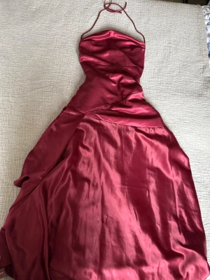 Vinröd maxiklänning - Snygg vinröd långklänning i satinliknande material med halterneck snörning och öppen rygg med snörningsdetaljer. Klänningen har en figurnära passform upptill och en slits . Perfekt för speciella tillfällen. Använd en gång, passar mig som är 165cm och har storlek s/m.
